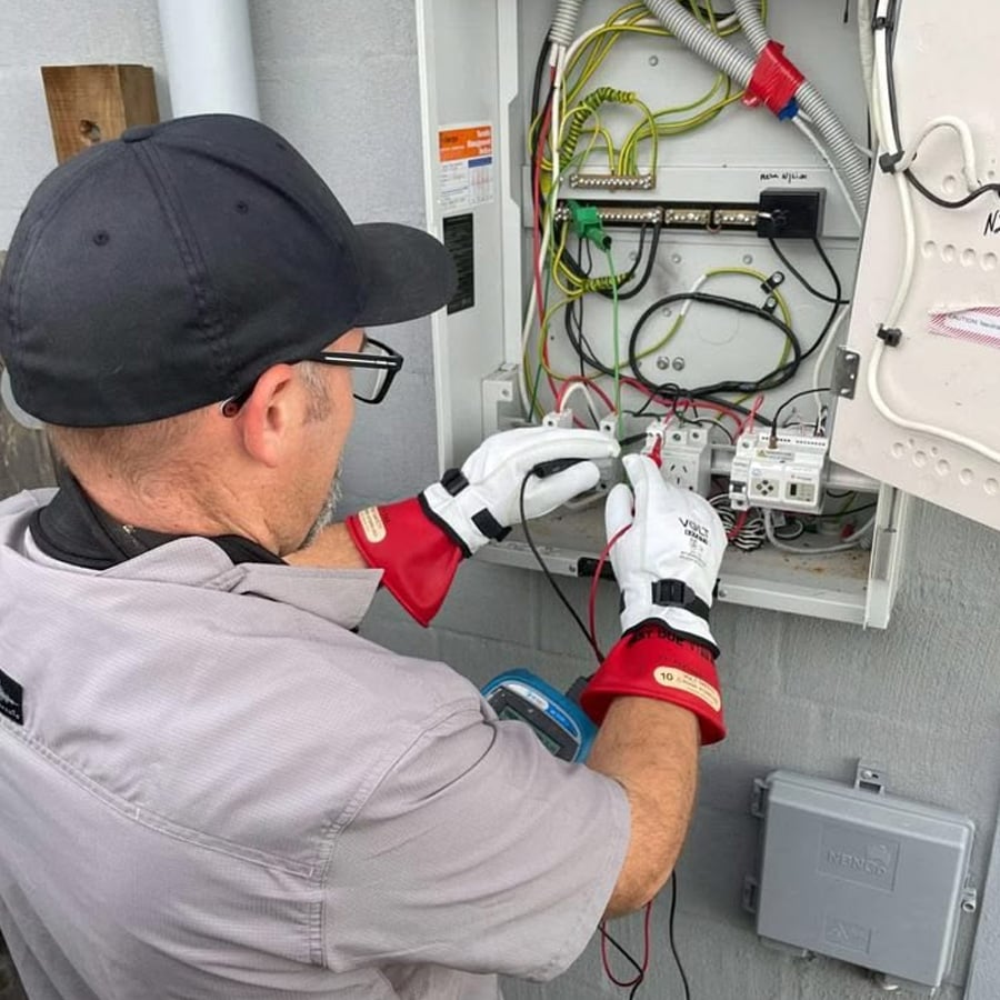 An electrician in safety gloves works on a control panel with various colored wires and electrical components installed on an exterior wall.