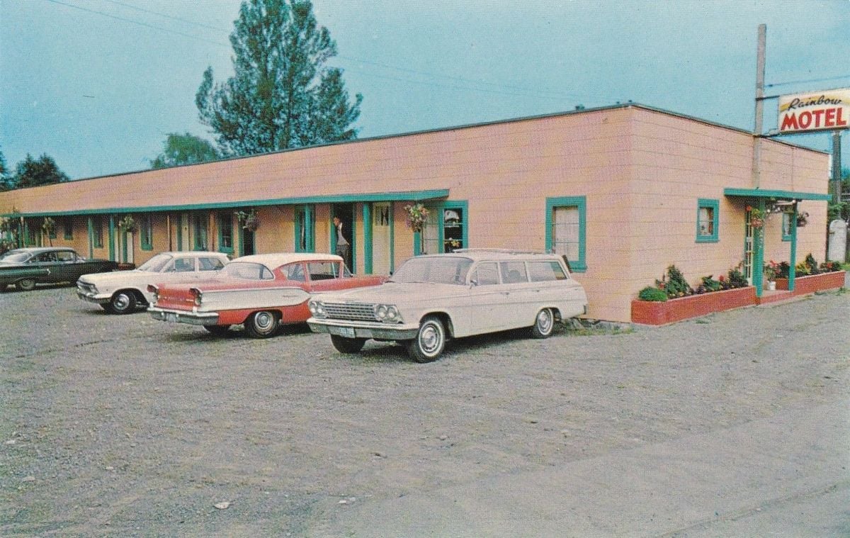 A vintage motel with a pink exterior and green trim, with several classic cars from the 1950s and 1960s parked in front. A sign reads "Rainbow Motel." The parking lot is gravel, and there are potted plants by the doors.