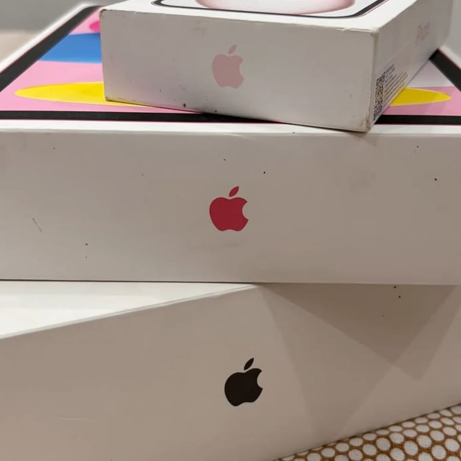 Three stacked Apple product boxes, each showing the Apple logo in different colors: red, black, and pink. The boxes are set against a multicolored background.