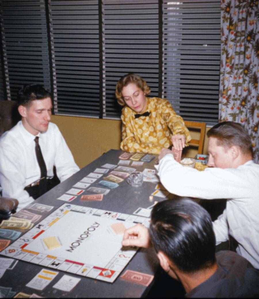 Four adults sit around a table playing Monopoly. The board and game pieces are visible, and they focus on the game. The room has blinds and patterned curtains in the background.