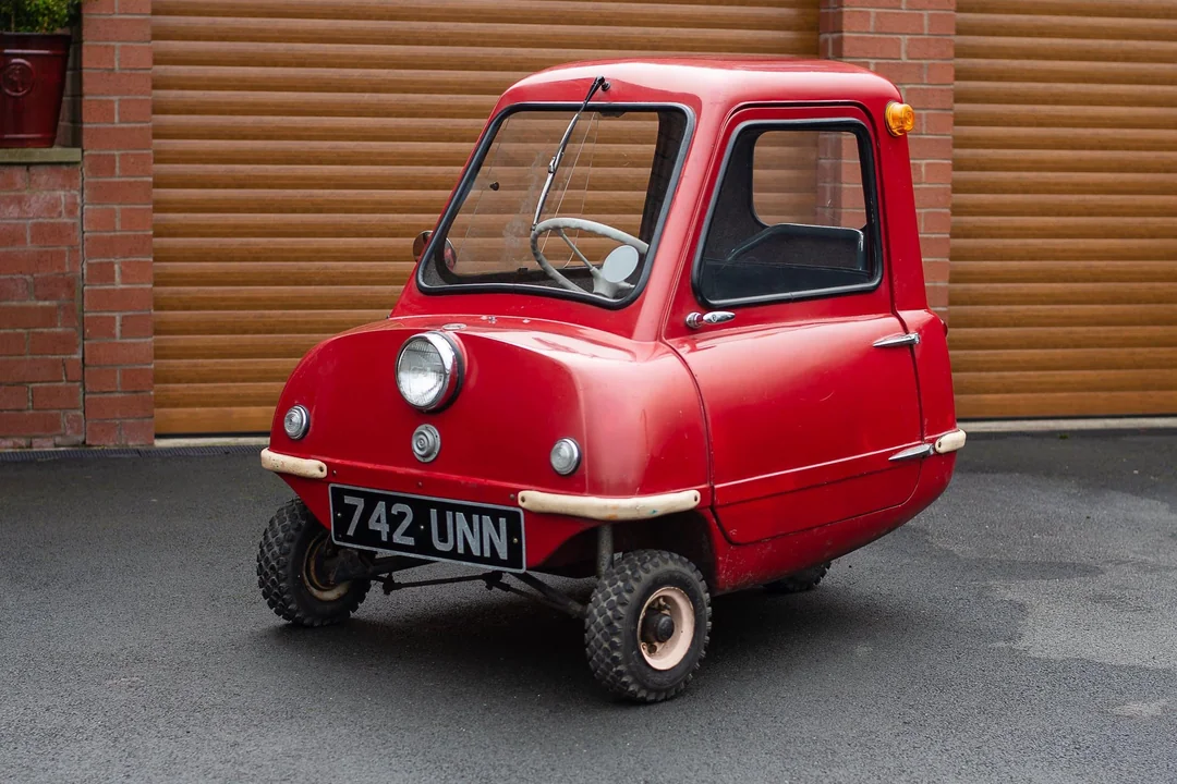 A small, red, three-wheeled car with a single headlight is parked on a paved driveway in front of brown garage doors and a brick wall. The license plate reads "742-UNN.
