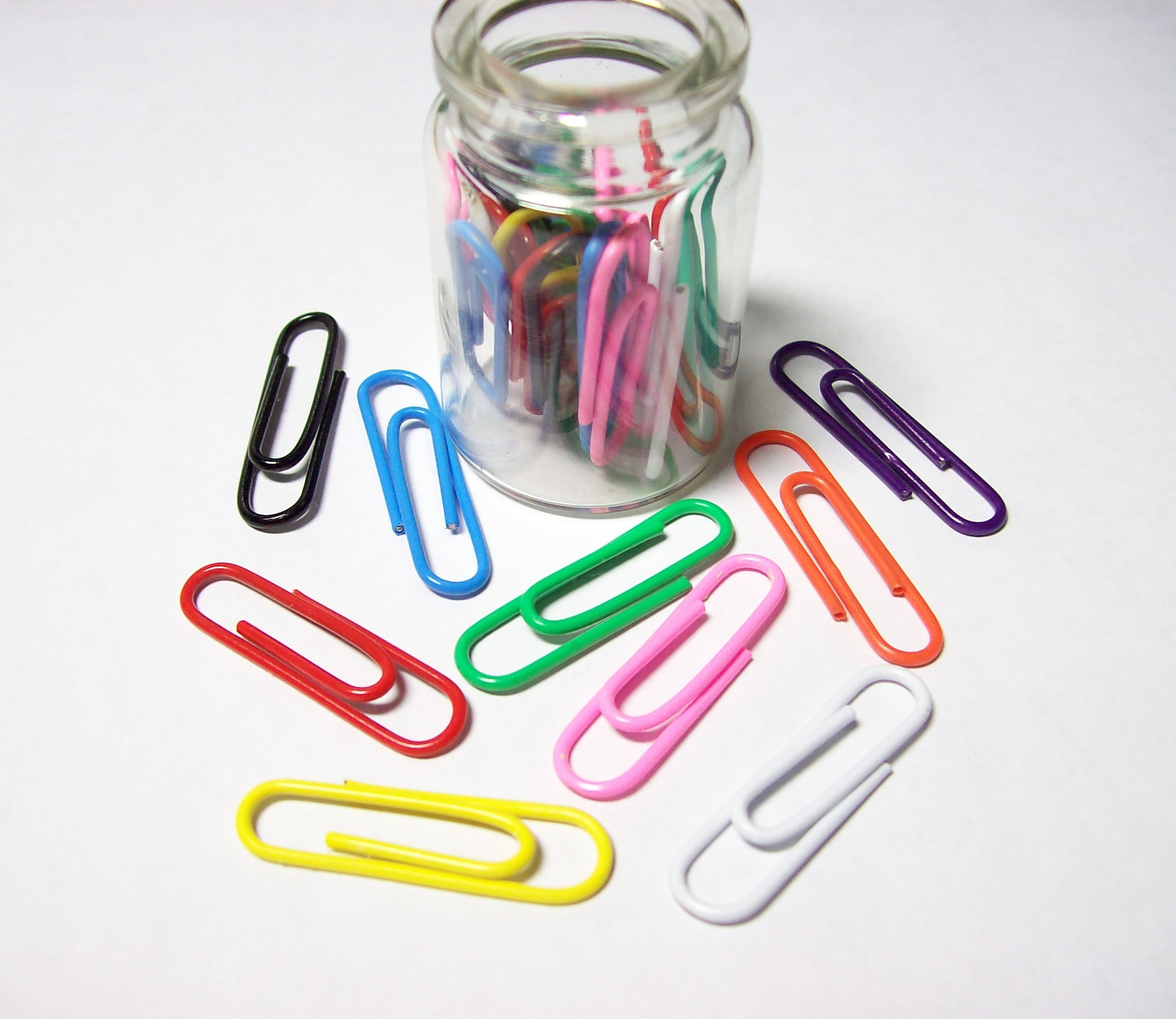 A small glass jar filled with colorful paperclips sits on a white surface. Several paperclips in various colors, including red, yellow, blue, and green, are scattered around the jar.