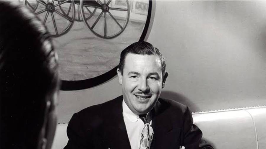 A man in a suit smiles while sitting on a sofa, with a circular painting of wagon wheels on the wall behind him. Another person is seen from behind in the foreground.