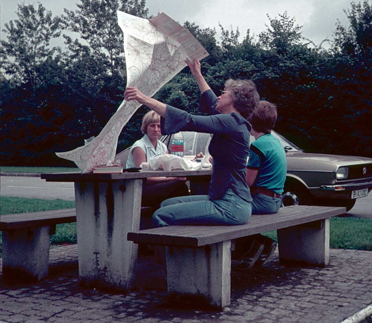 Two women and a man sit at a picnic table near a road, with one woman holding up a large, unfolded map. A green car is parked nearby, and there are trees in the background.