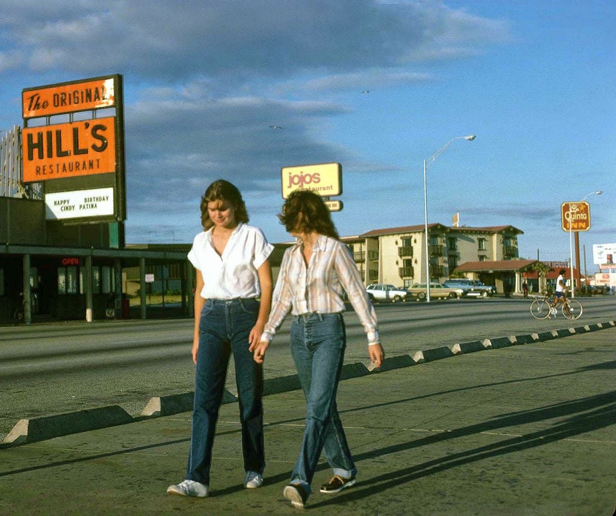 Two women in casual clothing walk together along a street with several restaurants in the background, including Hill’s Restaurant and Jojo’s. The sky is blue with some clouds, and a few people are seen farther back.