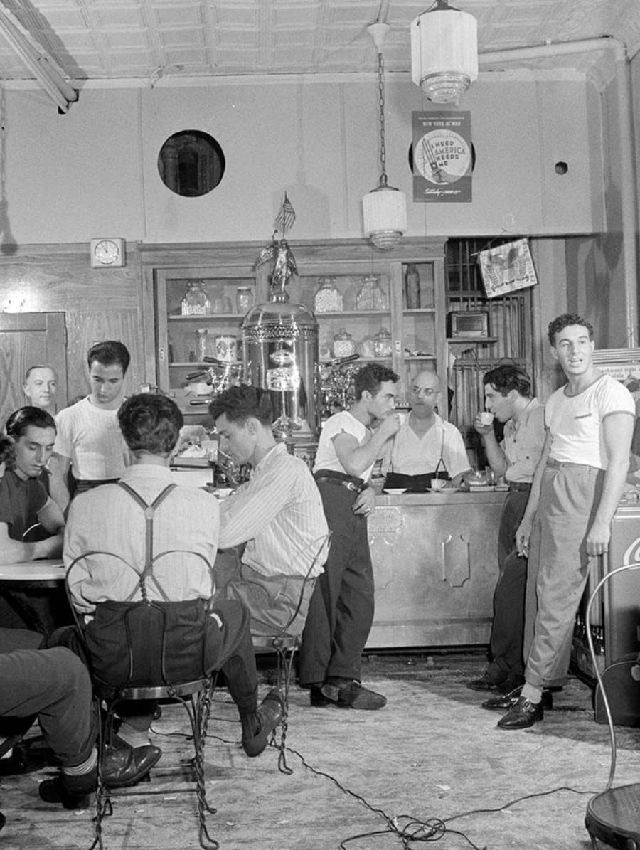 A group of men gather in a vintage café or bar, some sitting at tables, others standing near the counter, talking and relaxing. The room features retro furniture, hanging lights, and shelves with bottles and décor.