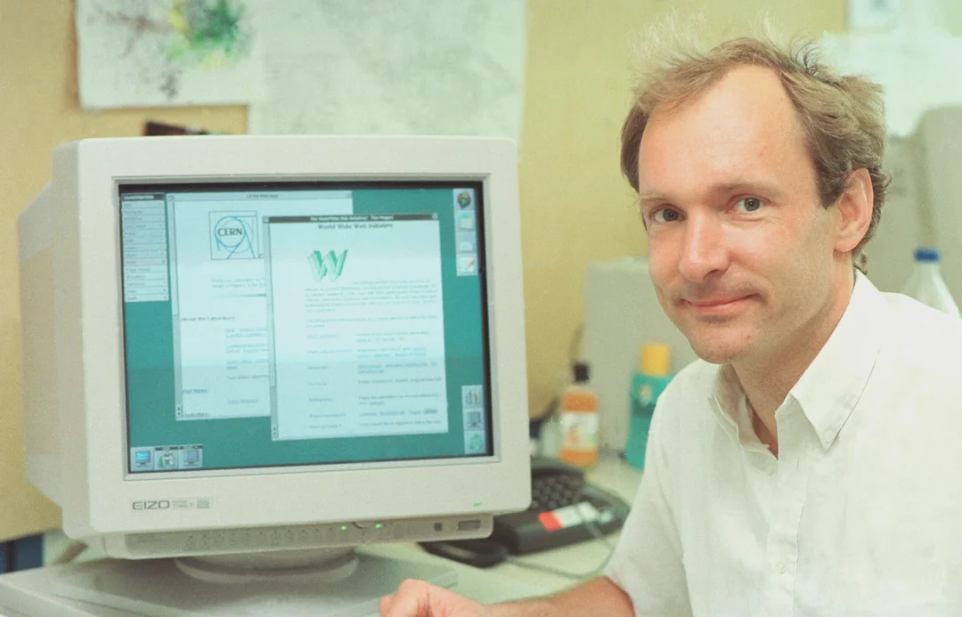 A man in a white shirt sits next to a vintage computer displaying an early web browser on the screen. The background shows office supplies and papers on a beige wall.