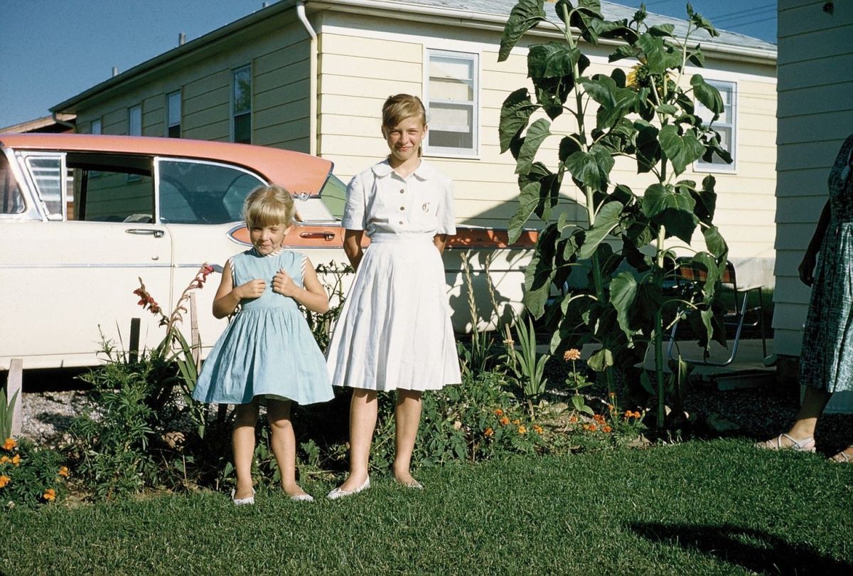 Two young girls in light blue dresses stand on a green lawn in front of a garden, with a vintage pink and white car and a light yellow house in the background. Tall sunflowers and other flowers are behind them.