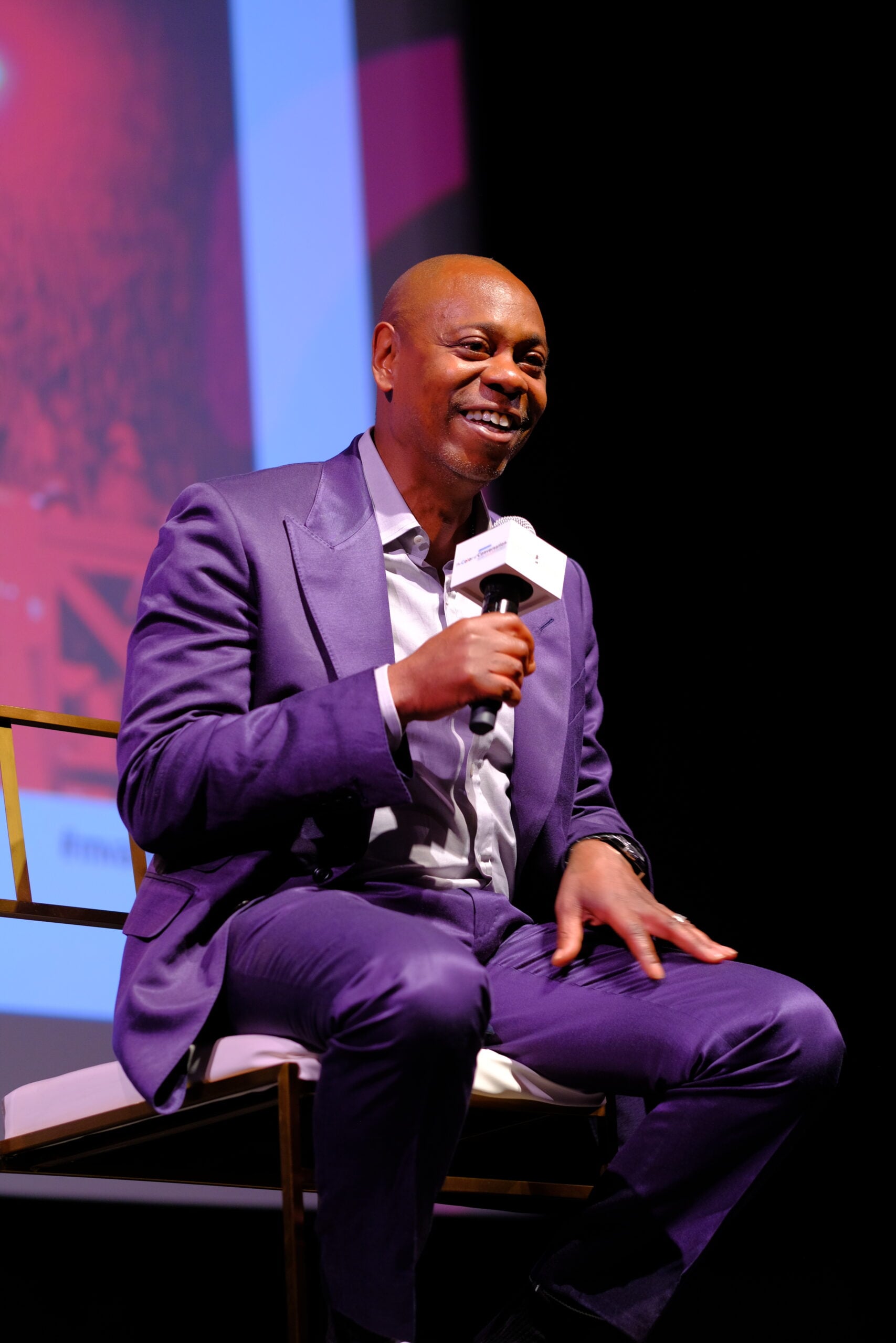 A man in a shiny purple suit sits on a chair, holding a microphone and smiling while speaking on stage, with a colorful screen in the background.