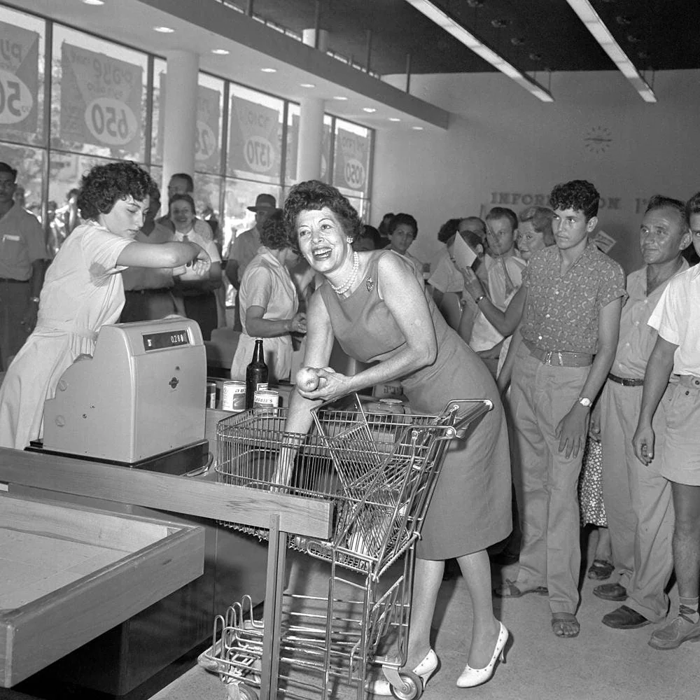 A woman smiles brightly at the checkout counter of a busy grocery store in the 1950s, while a cashier rings up her items and a line of customers waits behind her.