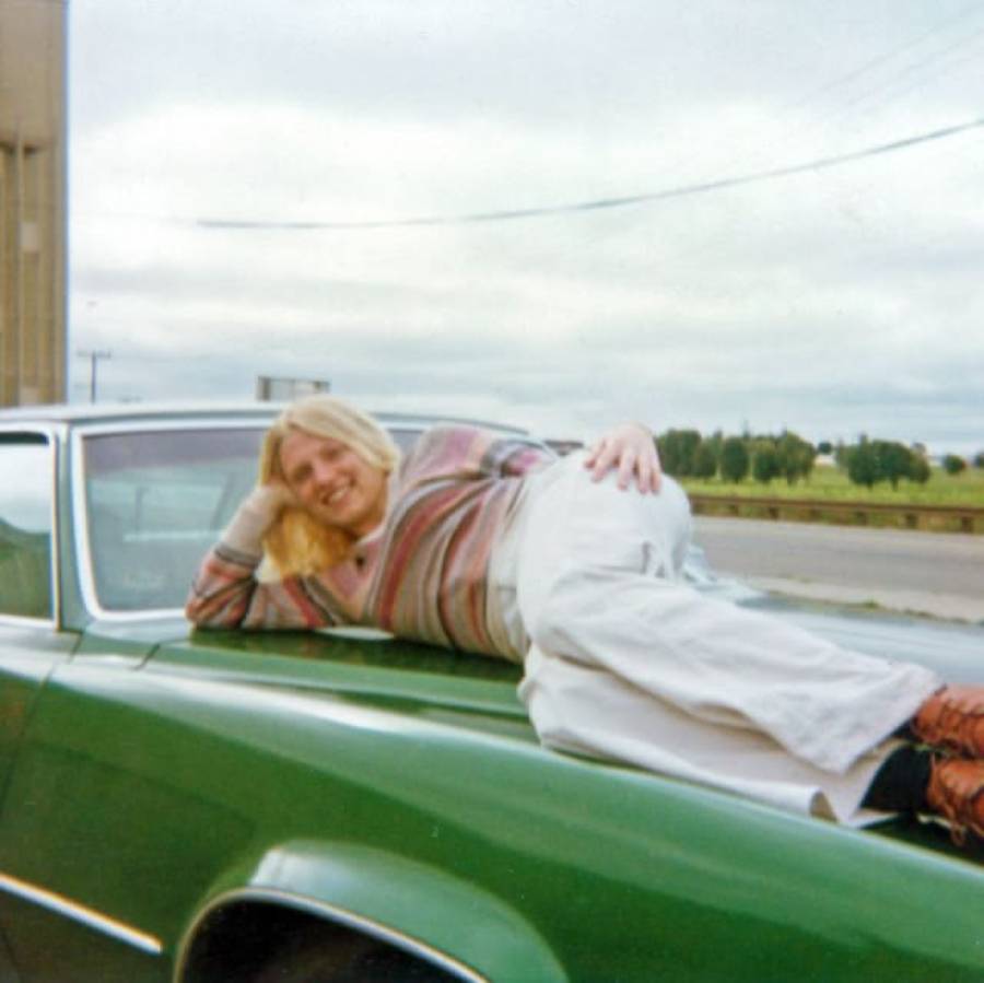 A person with long blond hair, wearing a striped sweater and light-colored pants, is smiling and reclining on the hood of a green car parked beside a road on a cloudy day.