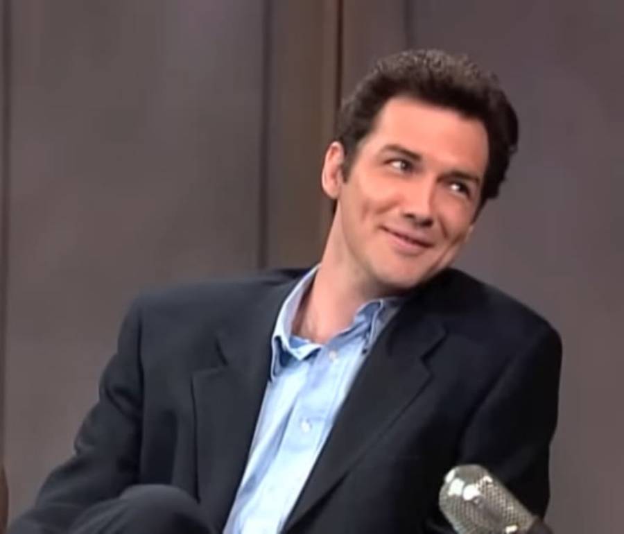A man in a dark suit and blue shirt sits and smiles while looking to the side on a talk show set, with a microphone visible in the foreground.