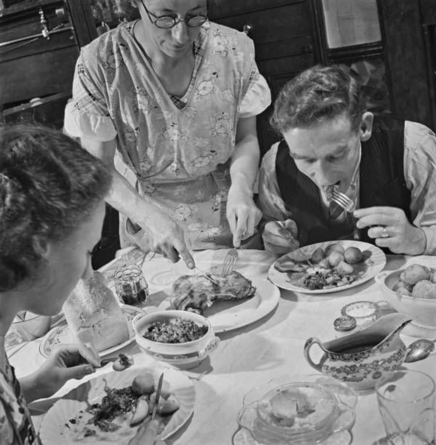 A woman serves food to two people at a dining table covered with plates of meat, potatoes, and vegetables. One man is eating hungrily while another woman watches. The scene appears to be from a past era.