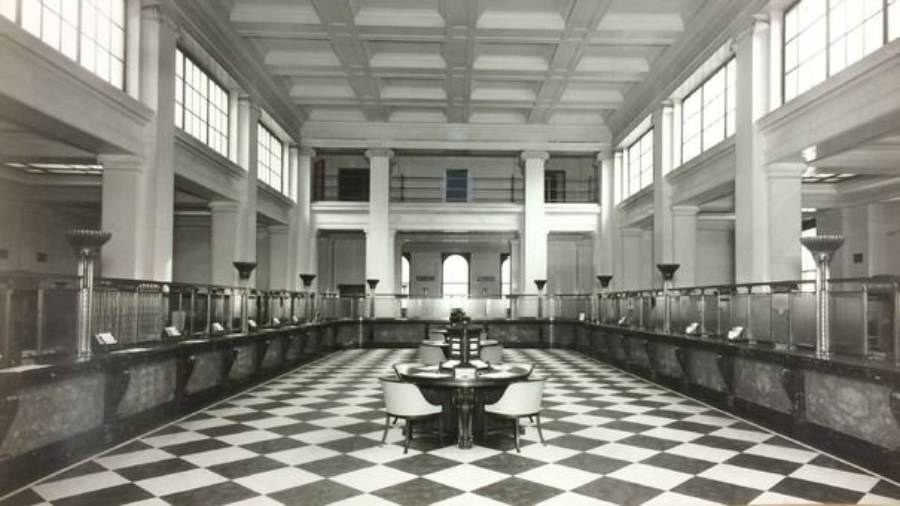 Black and white photo of a spacious, empty bank lobby with high ceilings, checkered tile floor, and teller counters lining the walls. Large windows let in natural light. Two round desks with chairs are in the center.