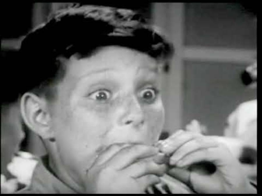 A surprised boy with wide eyes holds his hands up to his mouth as if eating or reacting in shock. The image is in black and white.