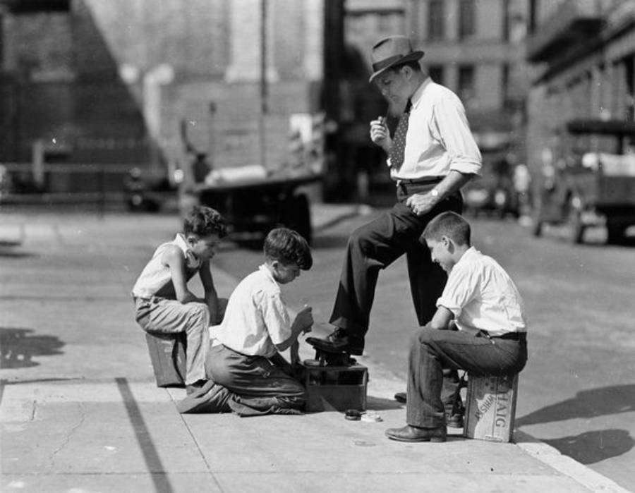 Three boys shine a man's shoe on a city sidewalk in what appears to be a historic, urban setting. The man stands with one foot raised, while the boys work with brushes and polish. Other buildings and cars are in the background.