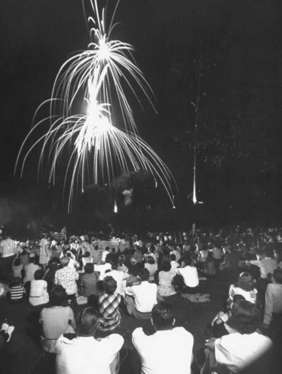 A large crowd sits outdoors at night watching bright fireworks burst in the sky above them. The scene is lively, with many people facing the display.