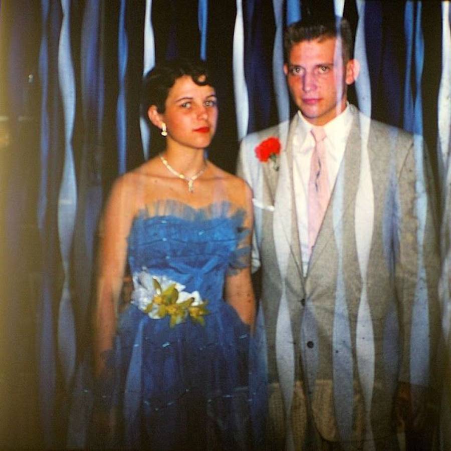 A young woman in a blue dress and a young man in a light suit with a red boutonniere pose together in front of blue and white streamers. The woman holds a yellow and white corsage.