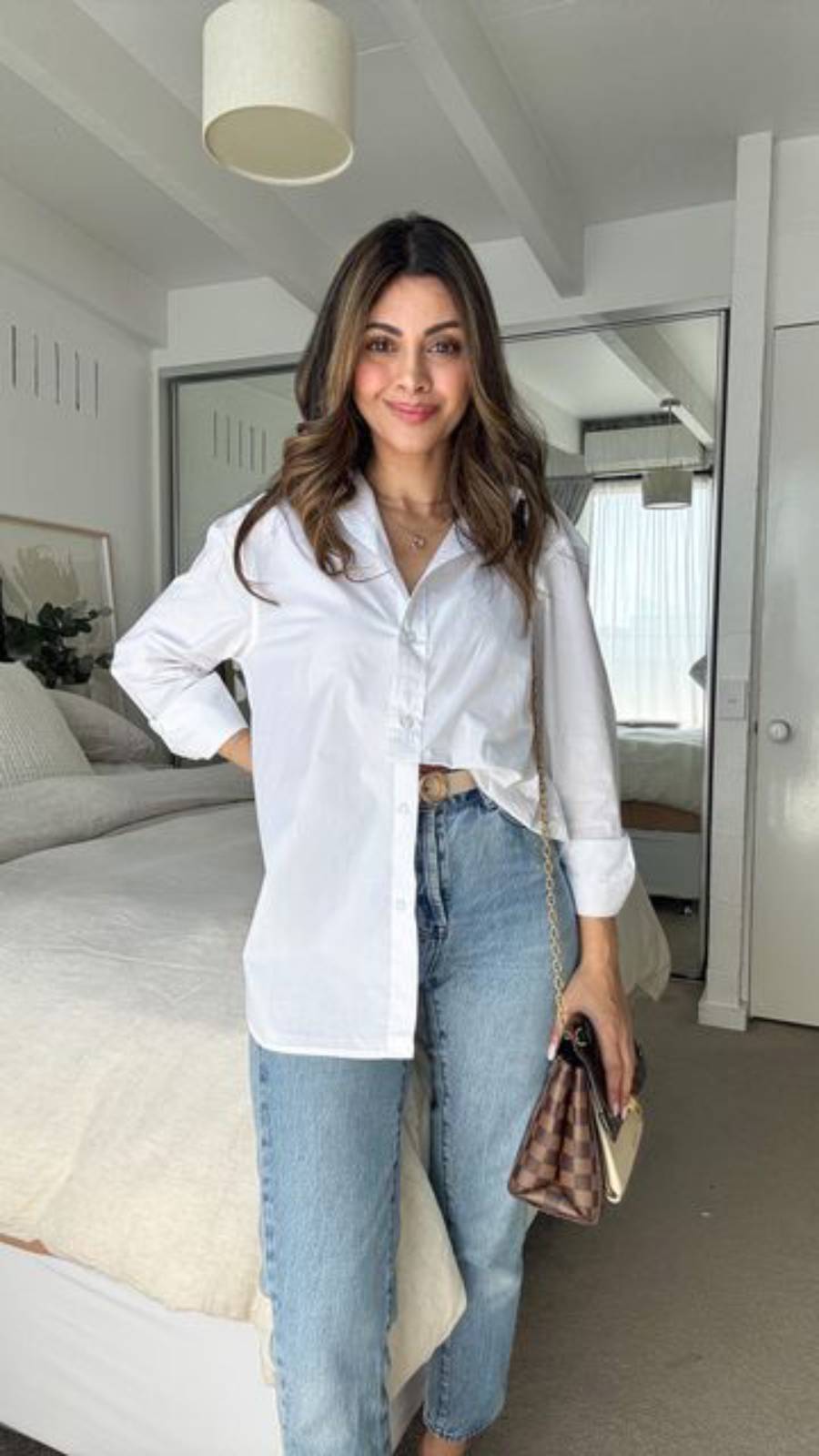 A woman stands in a bright bedroom wearing a white button-up shirt and blue jeans, holding a small purse. The room has white walls, a bed with light bedding, and a large mirror.