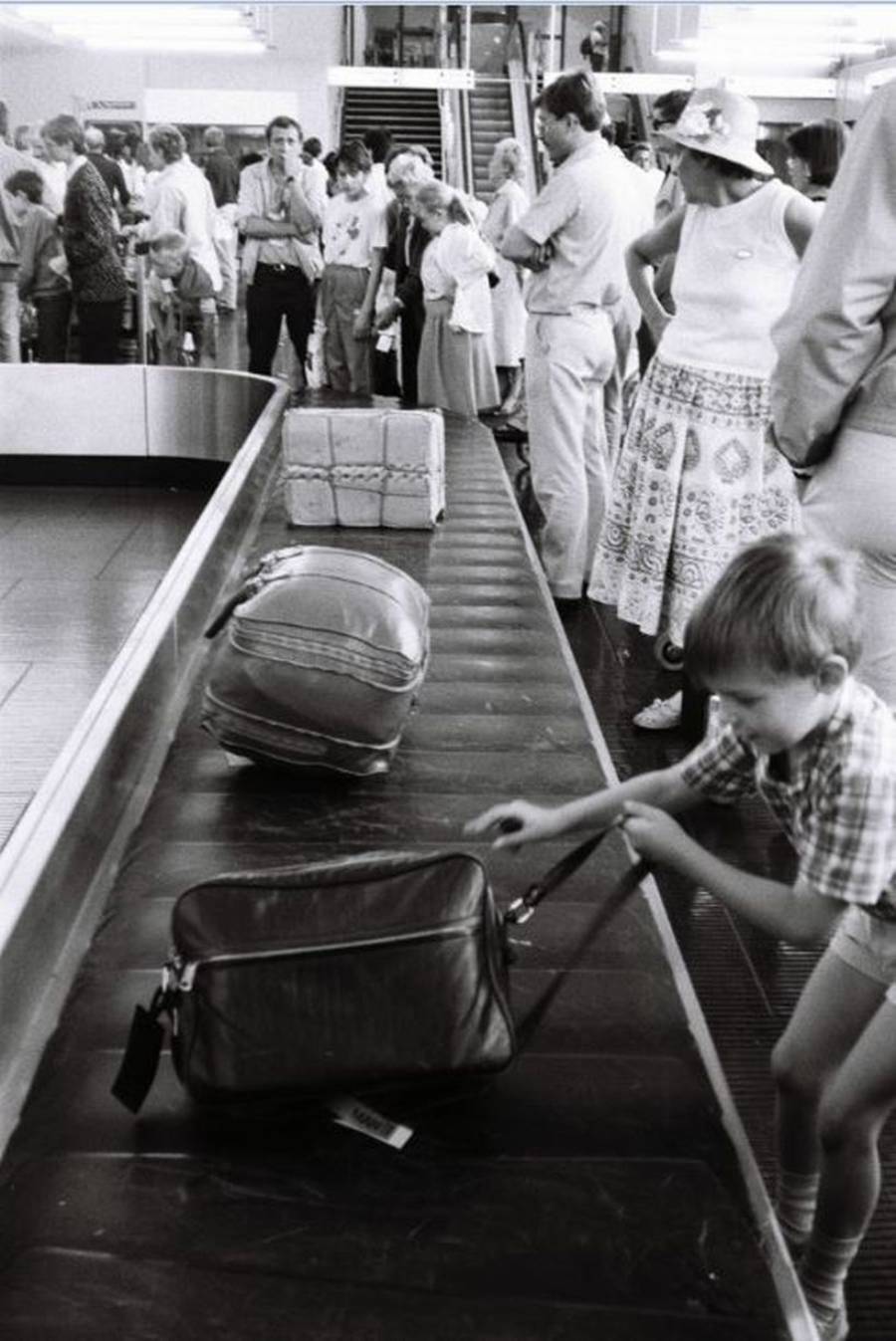 A black and white photo of a busy airport baggage claim; a young boy reaches for a suitcase on the conveyor belt while adults wait nearby. People in the background watch the scene and stand near stacked luggage.