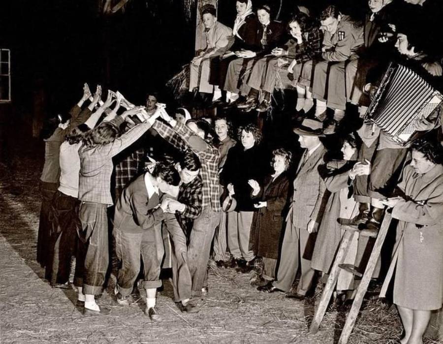 A group of people form a human tunnel while others walk through it; more people sit above, some watching, one playing an accordion.
