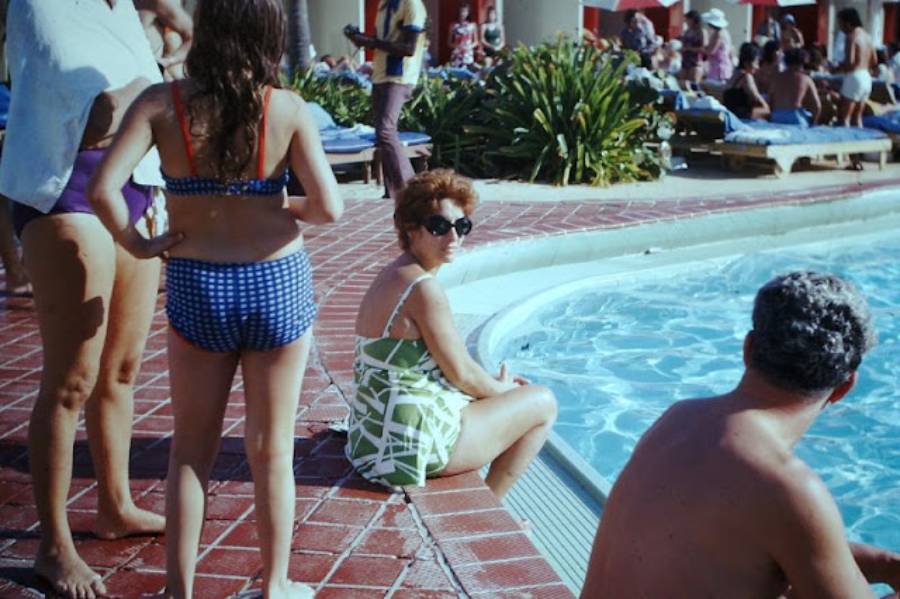 Several people relax by a pool. A woman in sunglasses sits at the pool’s edge, while a child in a blue swimsuit stands nearby. Other people lounge and walk in the background.