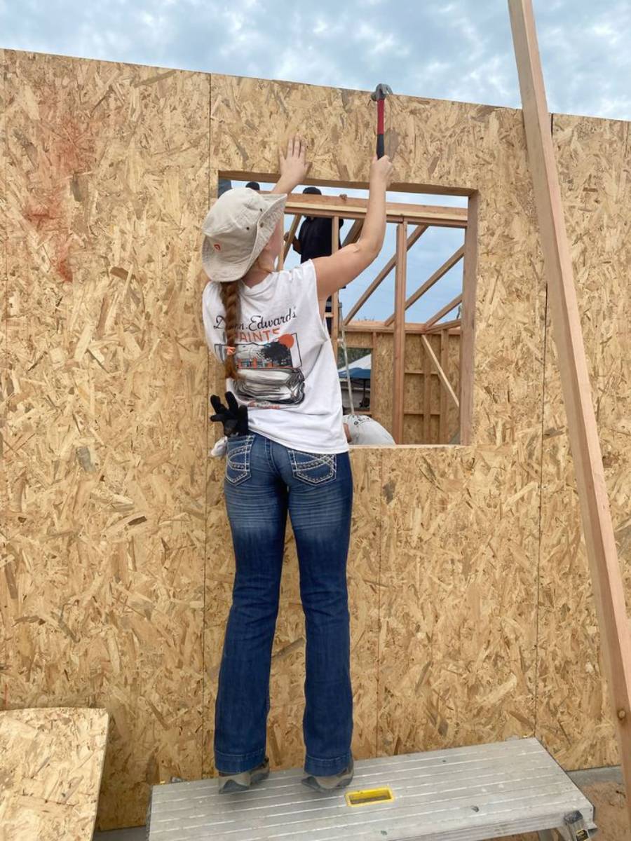 A person in jeans and a t-shirt uses a hammer to work on a wooden wall with a window opening at a construction site. They are standing on a platform and wearing a baseball cap.