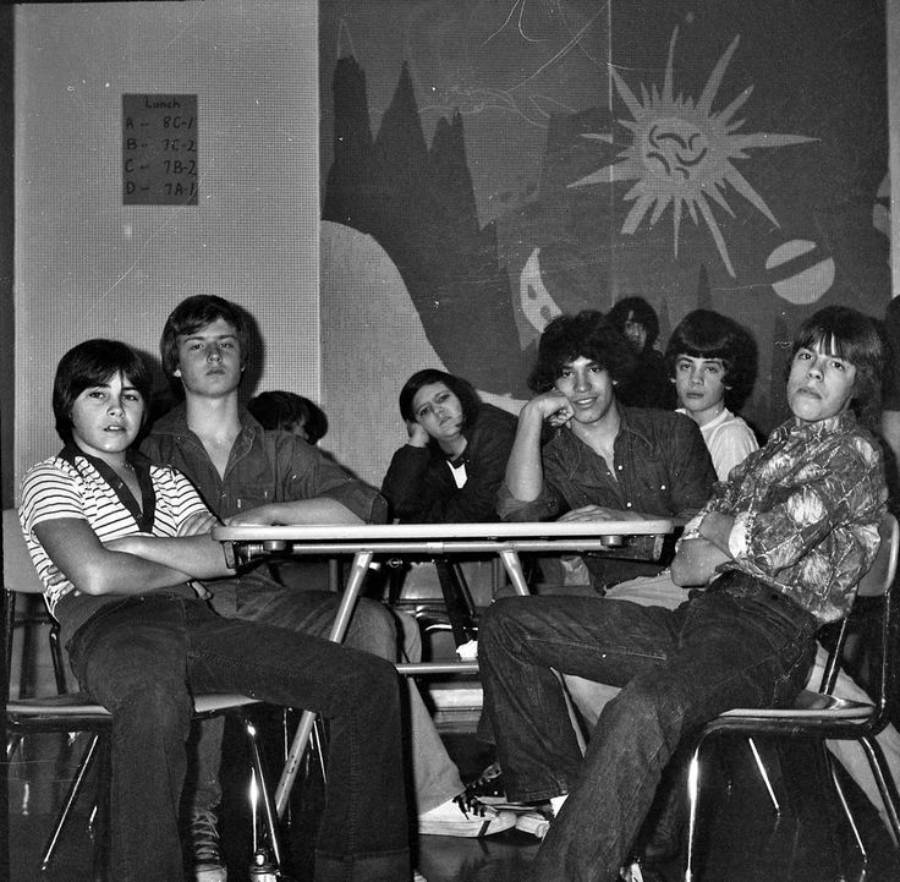Six teenagers sit around classroom desks, casually posing for a black and white photo. A painted mural with mountains, sun, and planets decorates the wall behind them. A lunch menu is posted to the left.