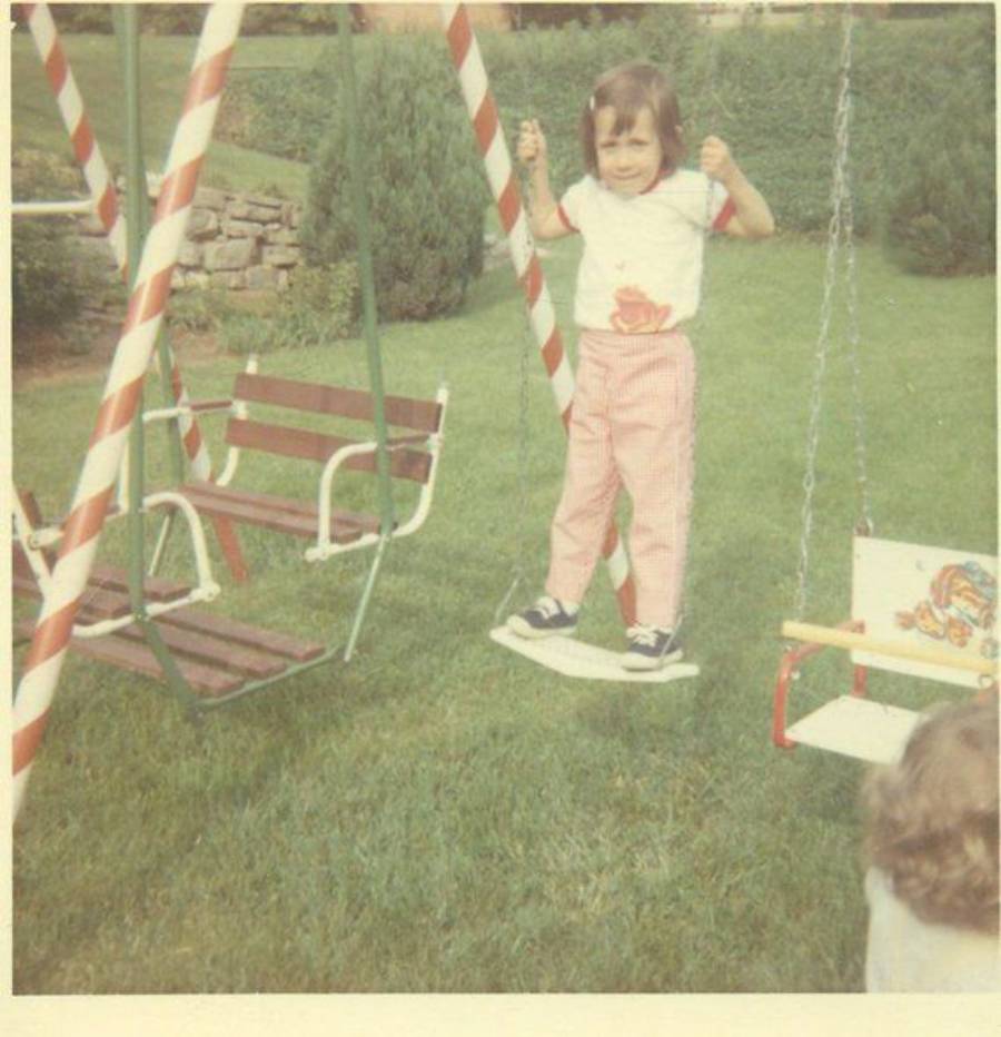 A young child stands on a swing in a grassy yard, holding the swing's ropes. Another swing and a small bench swing are nearby. Trees and bushes are in the background.