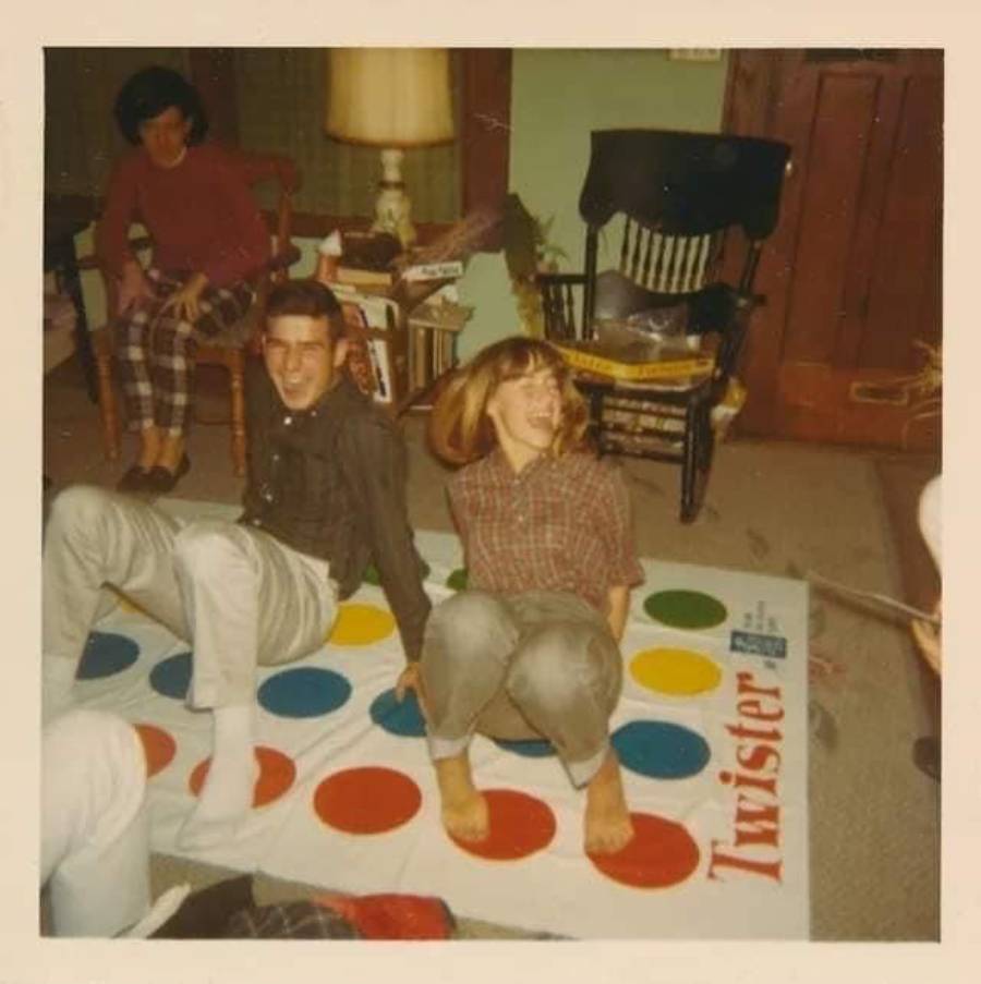 Two people laugh while sitting on a Twister game mat indoors, with another person watching from a chair. The room has a lamp, books, and a wooden rocking chair in the background.