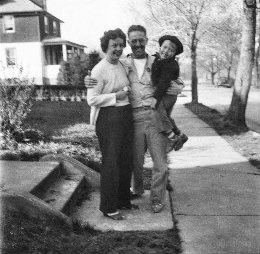 A smiling woman, man, and young boy pose together on a sidewalk near some steps and trees. The man hugs the woman and holds the boy in his arms. A house is visible in the background on a sunny day.