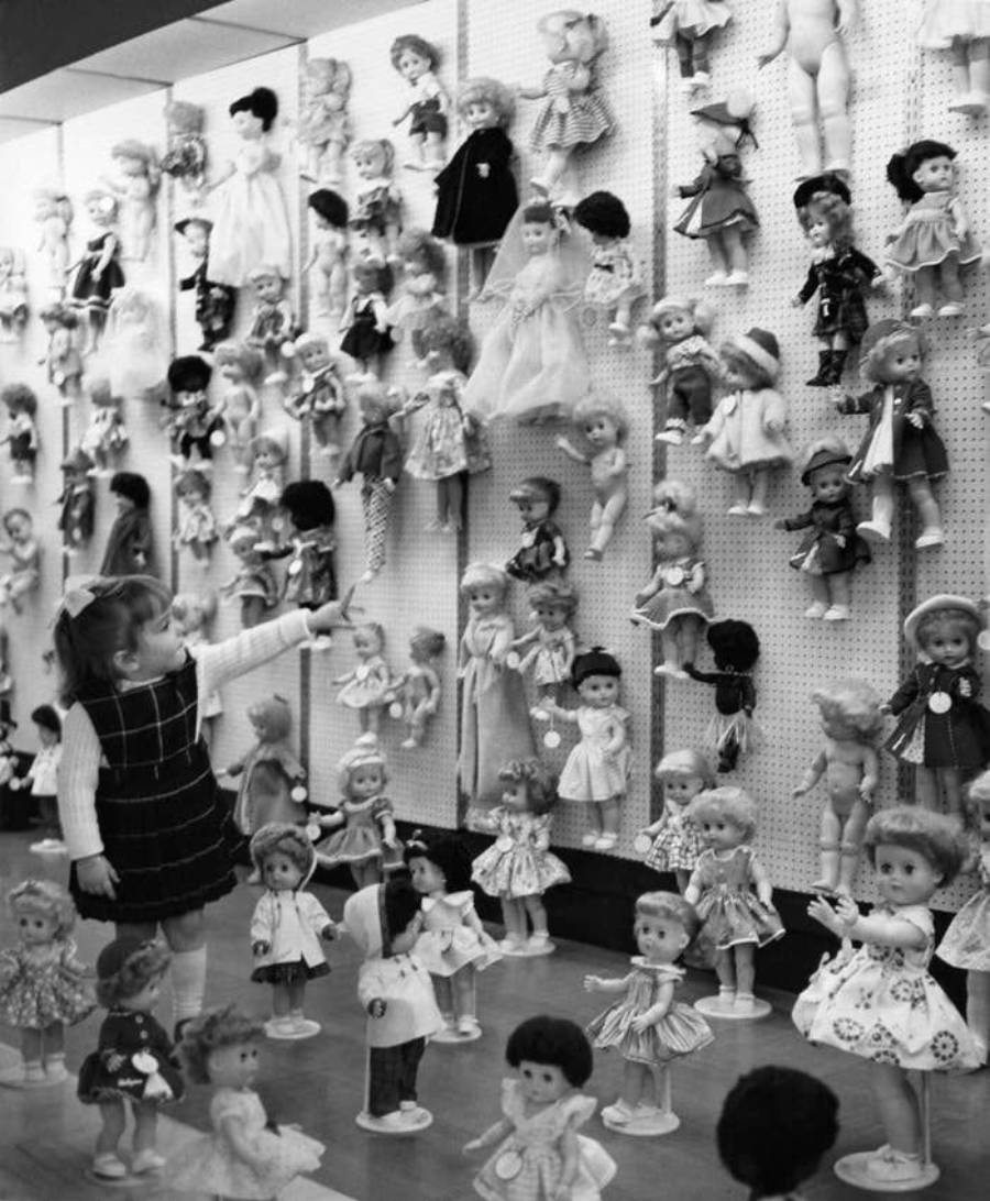 A young girl points at a wall filled with various dolls in dresses, with more dolls arranged on stands at her feet in a toy store or museum setting.