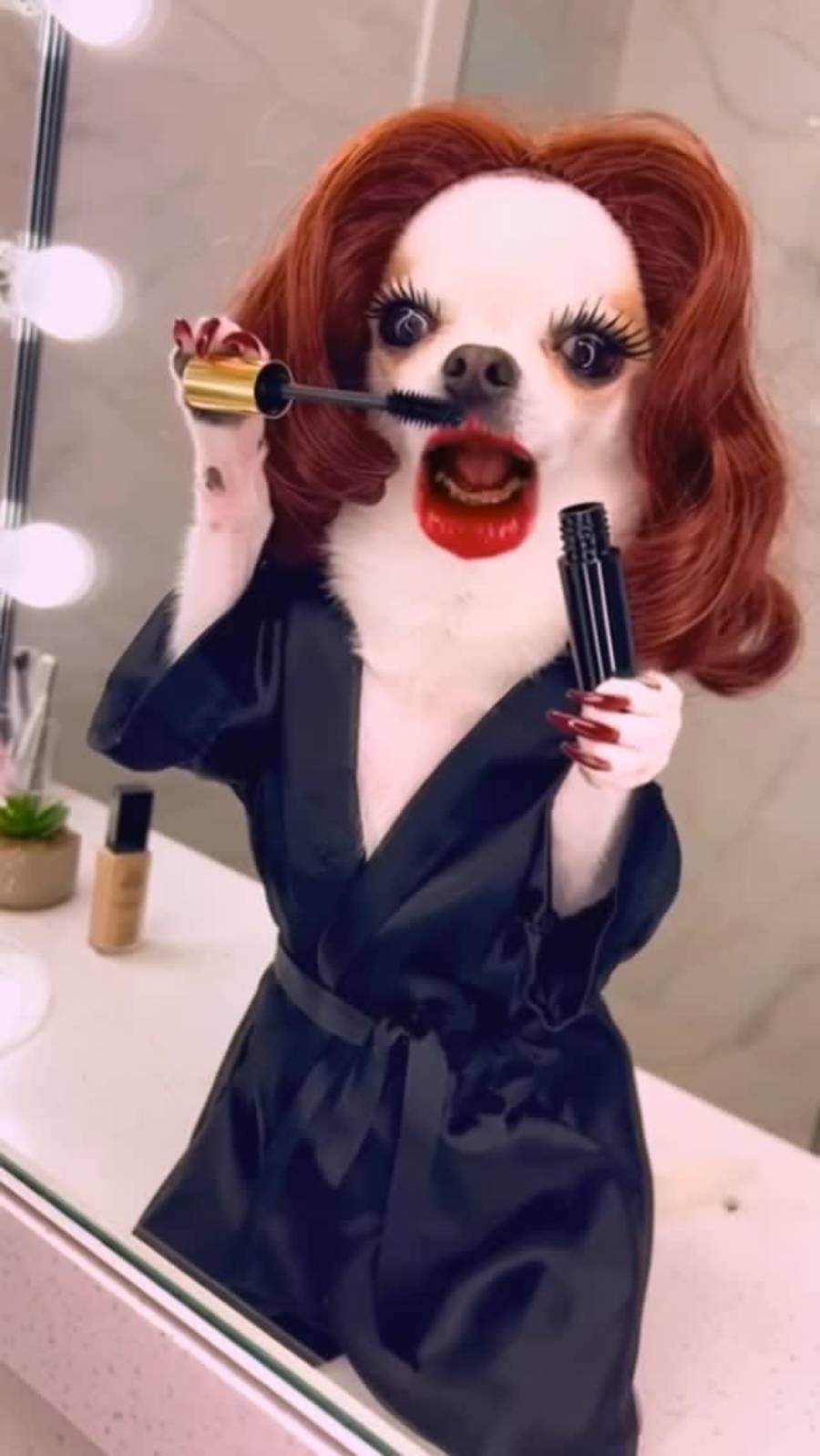 A Chihuahua wearing a red wig, black silk robe, and dramatic makeup—including long lashes, red lipstick, and long nails—holding a mascara wand in a bathroom setting with vanity lights.