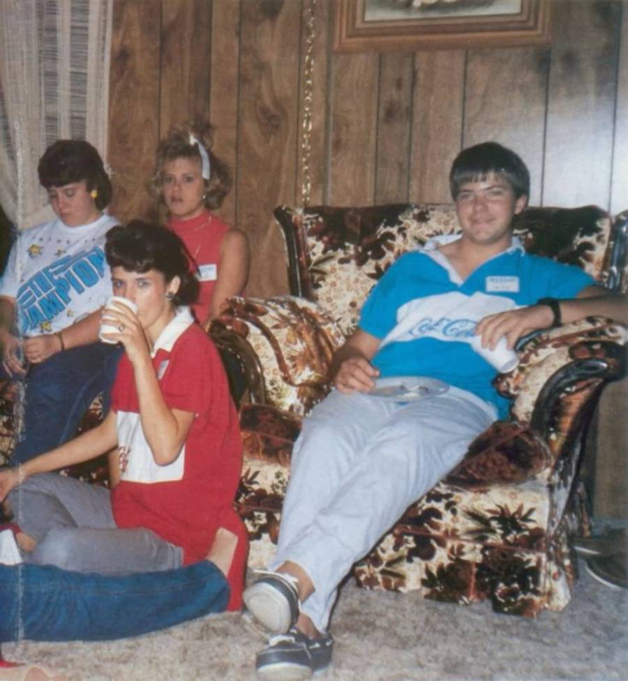 Four people sit in a living room with wood-paneled walls and floral furniture. Three are on a couch; one on an armchair. The group appears relaxed, with one person drinking from a cup and another holding a rolled-up T-shirt.