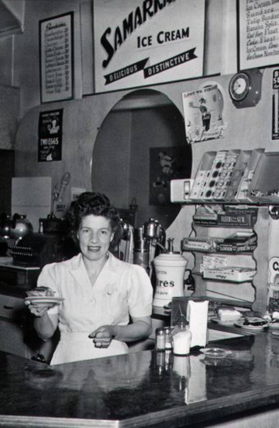 A woman in a white uniform stands behind a diner counter, holding a plate of food. Behind her are shelves with eggs, canned goods, and a sign advertising Samaritan Ice Cream. The setting appears to be a vintage diner.