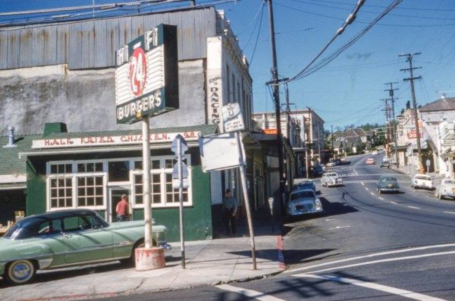 A vintage street scene features classic cars, a green and white diner with a sign reading "HALF FRIED CHICKEN," and a tall sign for "Hi Fi Burgers." Power lines and shops line a sloping road under a clear blue sky.