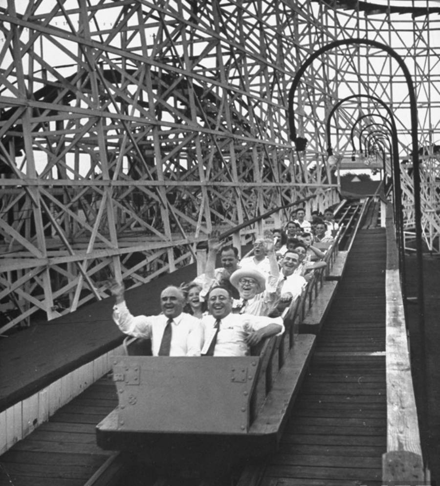 A group of people, dressed in vintage clothing, ride a wooden roller coaster, smiling and waving their arms as the coaster descends. The ride's intricate wooden supports are visible in the background.