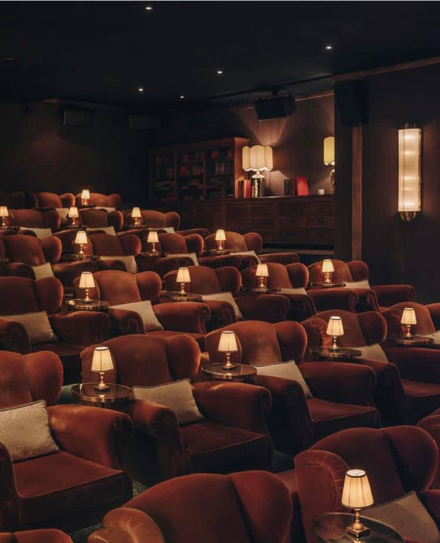 A cozy, dimly lit theater with rows of plush brown armchairs, each paired with a small table and a lamp, creating an intimate and inviting atmosphere.