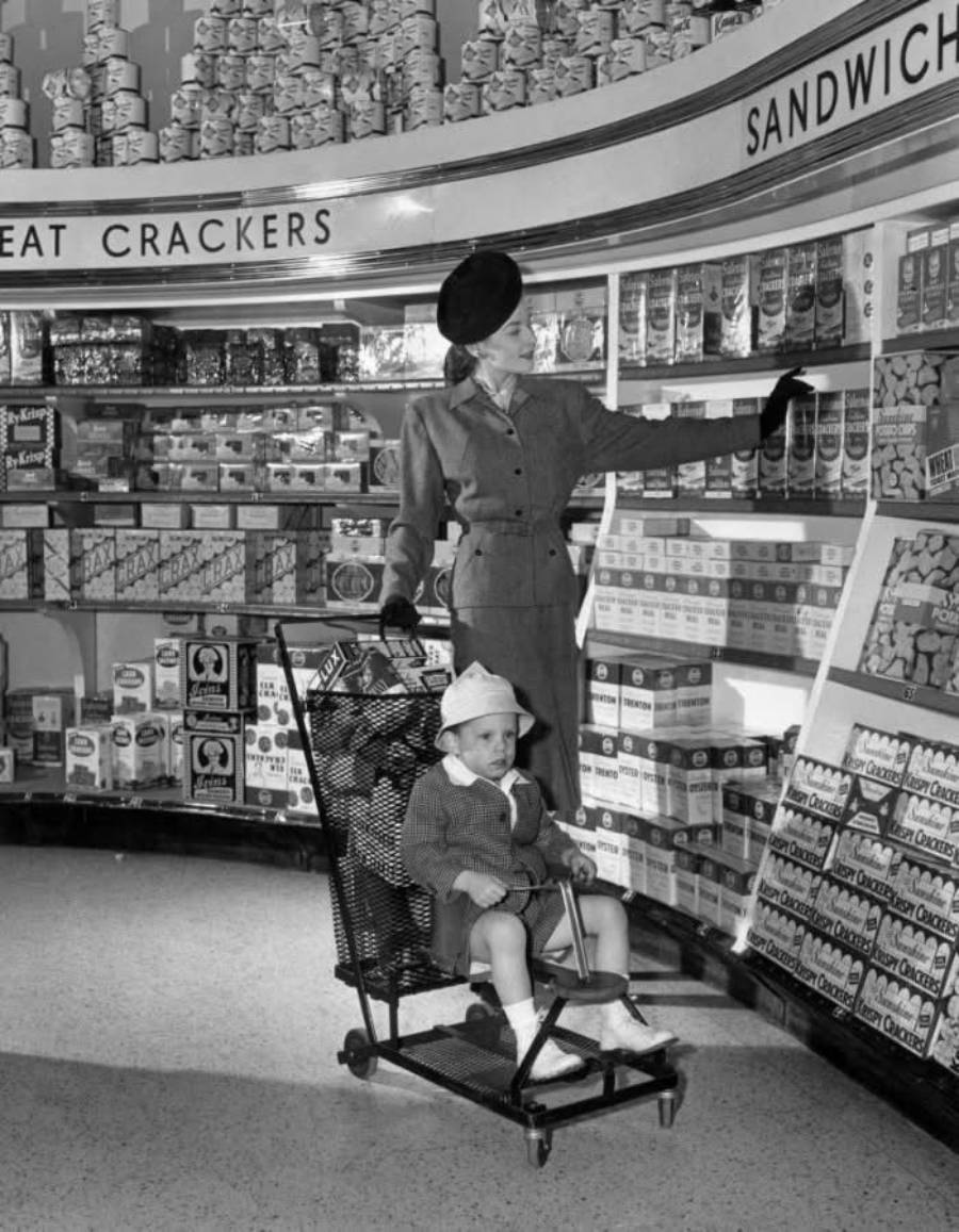 A woman in 1950s attire shops for crackers in a grocery store, pushing a shopping cart with a young child sitting in the front seat. Shelves are stocked with various boxes and tins of crackers and sandwiches.