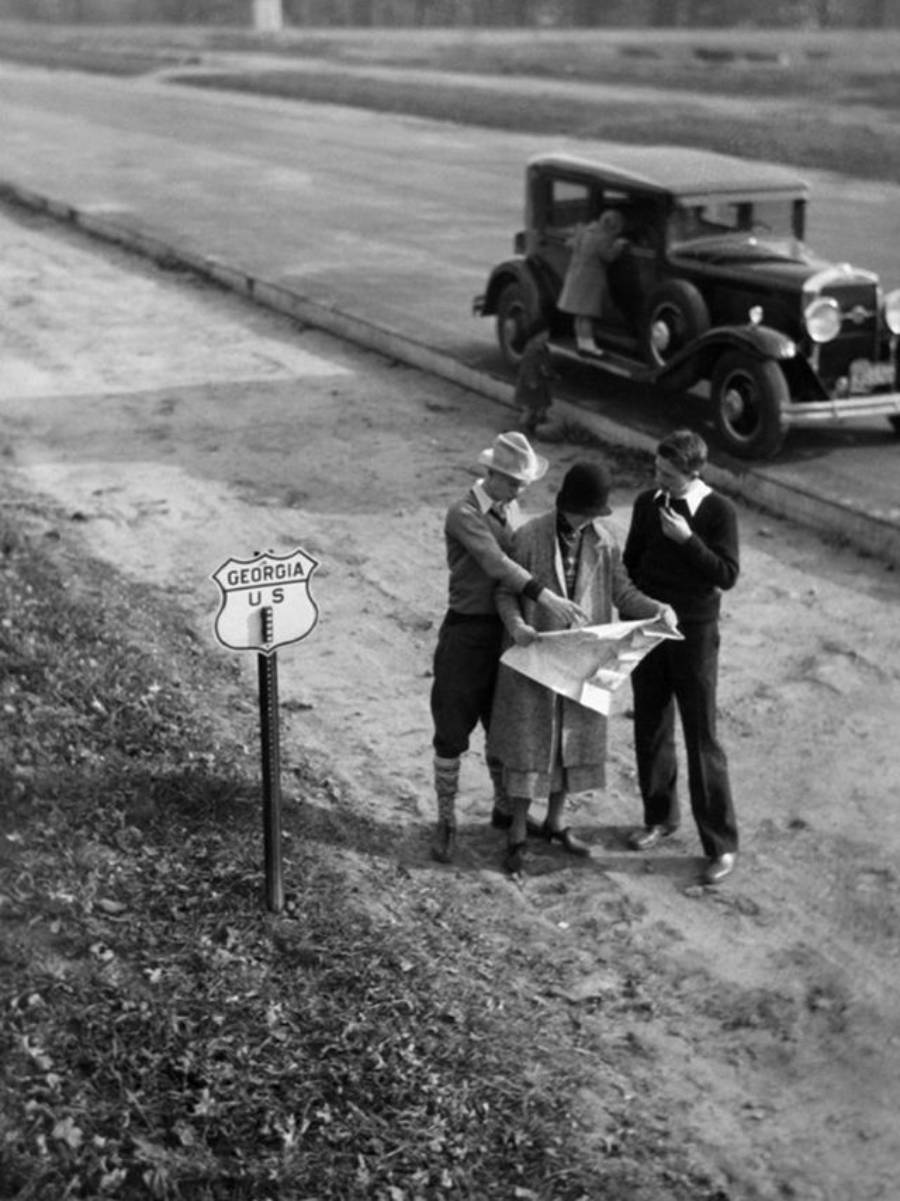 Three people stand on a roadside near a "Georgia US" highway sign, looking at a map. An old-fashioned car is parked nearby, with a person leaning inside. The scene appears to be from the early 20th century.