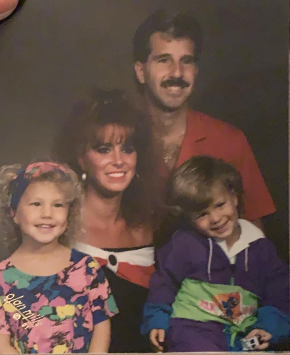 A smiling family of four poses for a portrait; two adults in bright clothing stand behind two children who are also smiling and wearing colorful outfits.