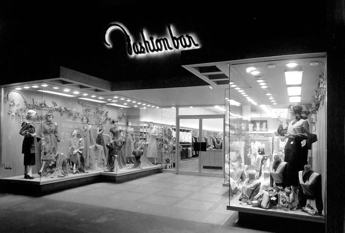 Black and white photo of a vintage clothing store called "Fashion Bar," with mannequins dressed in various outfits displayed in large front windows, and illuminated signage above the entrance at night.