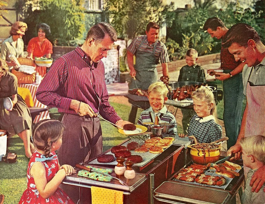 A group of adults and children gather outdoors for a barbecue. One man serves hamburgers while others grill and chat. Children wait in line, and picnic tables with food and drinks are visible in a sunny, backyard setting.