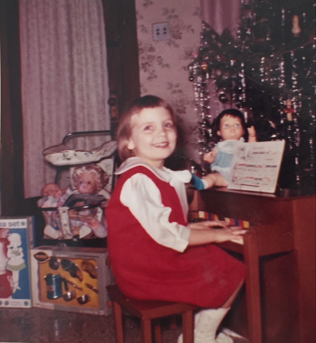A young girl in a red dress smiles while playing a small piano near a Christmas tree, surrounded by dolls and toys in a vintage living room.