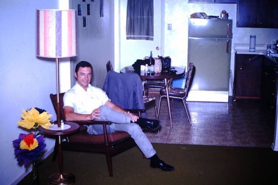 A man sits on a chair in a modest living room with a striped lamp, large colorful paper flowers, and a small kitchen and dining area visible in the background.