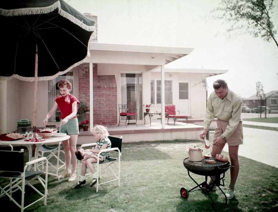 A woman prepares food at a table, a young girl sits with a doll, and a man grills outside a mid-century suburban home on a sunny day.