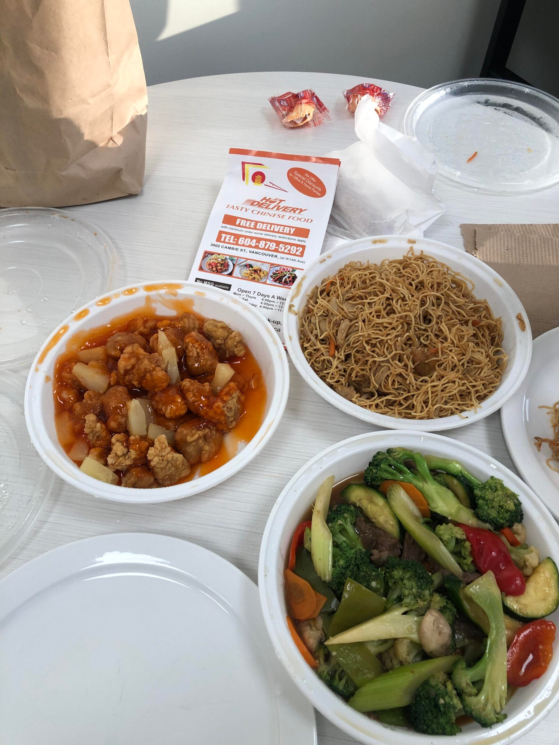 Three takeout dishes on a table: sweet and sour chicken, lo mein noodles, and mixed vegetables. A restaurant menu, fortune cookies, brown bag, and empty plates are also visible. Bright sunlight shines on the food.