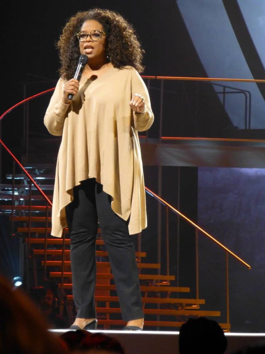 A woman stands on stage holding a microphone, wearing glasses, a tan asymmetrical top, black pants, and heels. Stairs and stage lights are visible in the background.