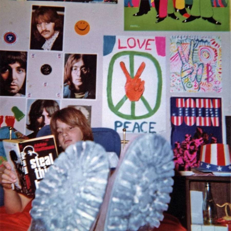 A person lounges with feet up, reading "Steal This Book" in a colorful room decorated with posters of The Beatles, peace signs, and 1960s-style artwork. The scene has a vibrant, retro and counterculture vibe.