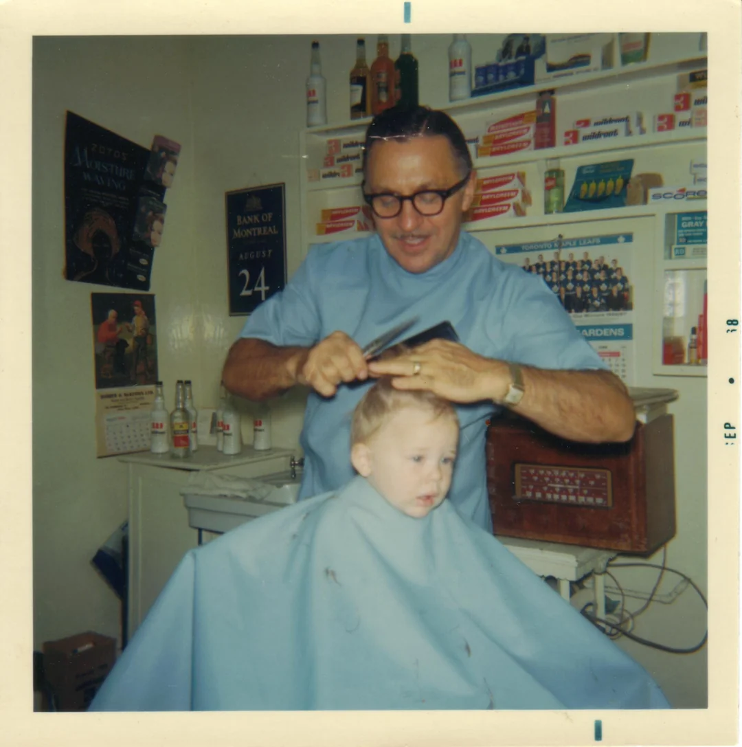 A barber in glasses cuts a young child's hair in a vintage barbershop, with shelves of bottles and boxes in the background. Both wear blue capes. The photo is dated September 1968.